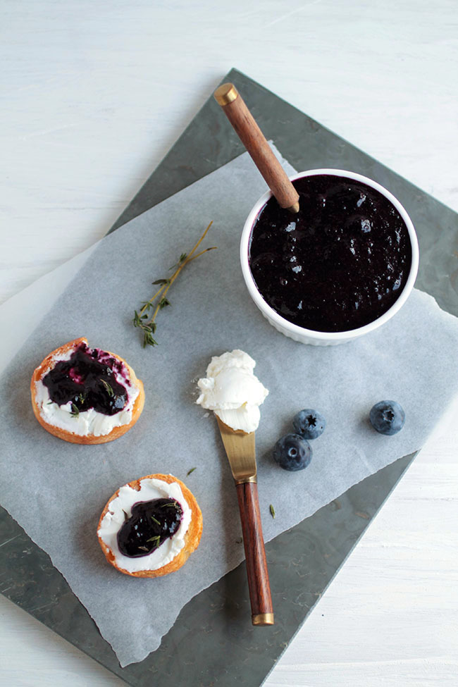 Slate platter with a bowl of blueberry jam, several crostini, and a knife holding a scoop of goat cheese.