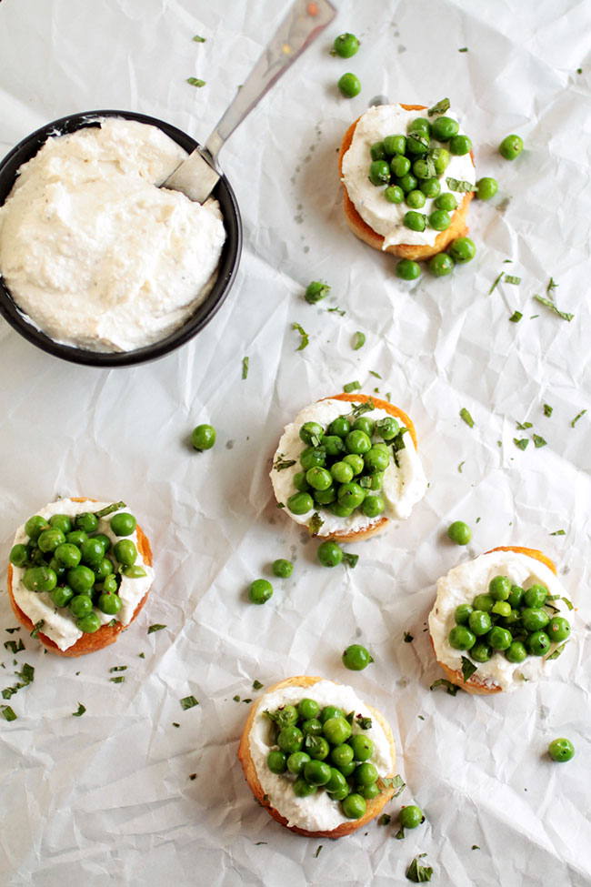 Overhead view of pea and mint crostini on a white surface, next to a bowl of whipped feta cheese.