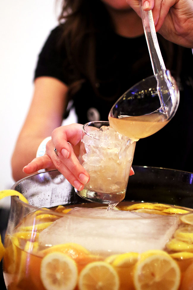 Bartender pouring cocktail into a small glass filled with ice, using a clear plastic ladle.