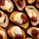 Multiple crostini on a dark sheet pan, each topped with a piece of melty brie cheese and a dollop of dark brown apple butter.