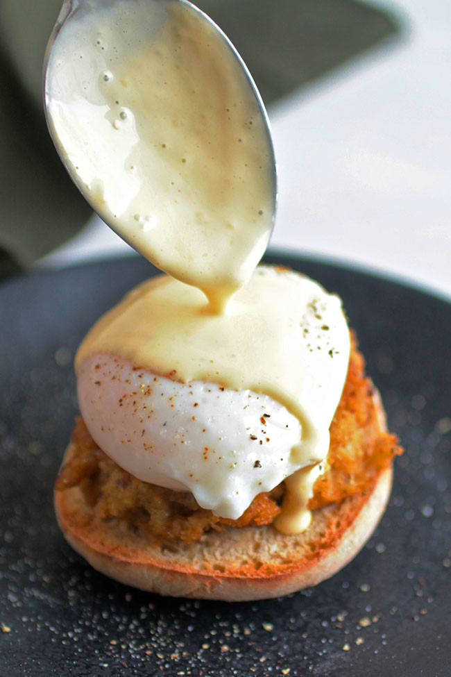 Spoon drizzling hollandaise sauce over eggs benedict.