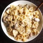 overhead photo of a white bowl filled with bowtie pasta and mushroom sauce on a dark brown table