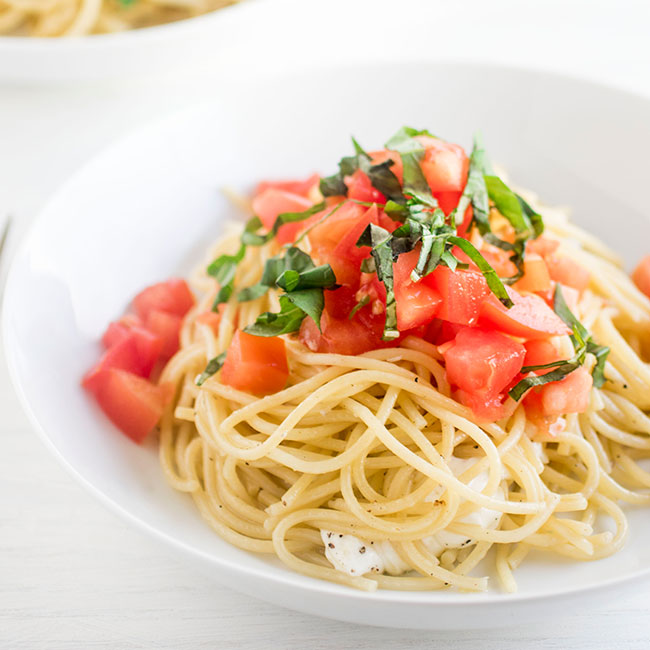 Spaghetti in a shallow white bowl topped with chopped tomatoes and sliced basil.