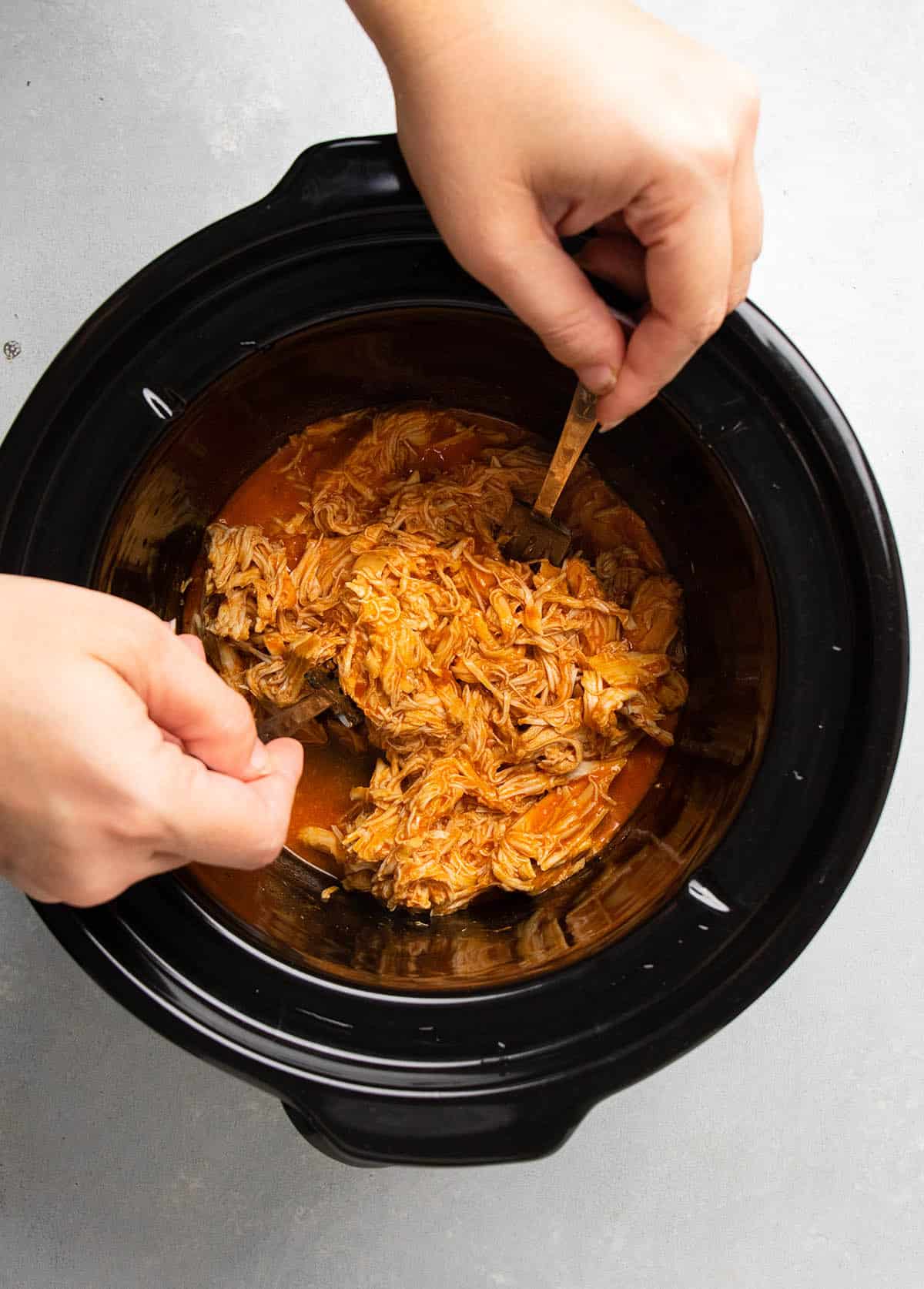 Two forks shredding buffalo chicken in a black slow cooker.