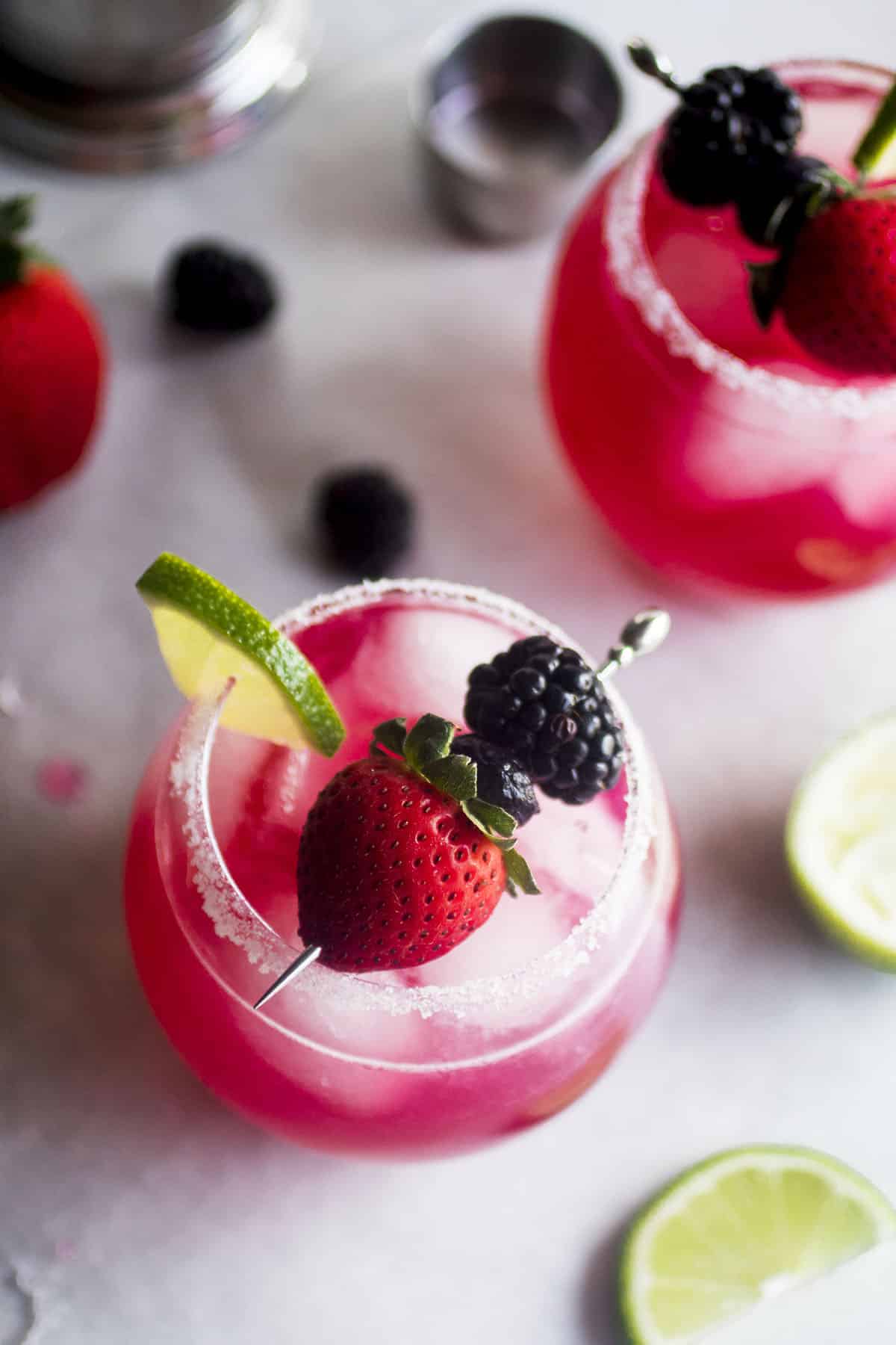 Two berry margaritas garnished with fresh fruit and lime wedges.