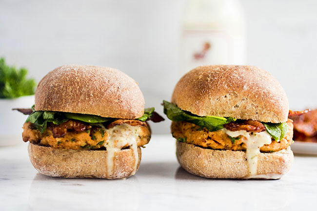 Two salmon burgers with tartar sauce dripping down the sides on a white background.