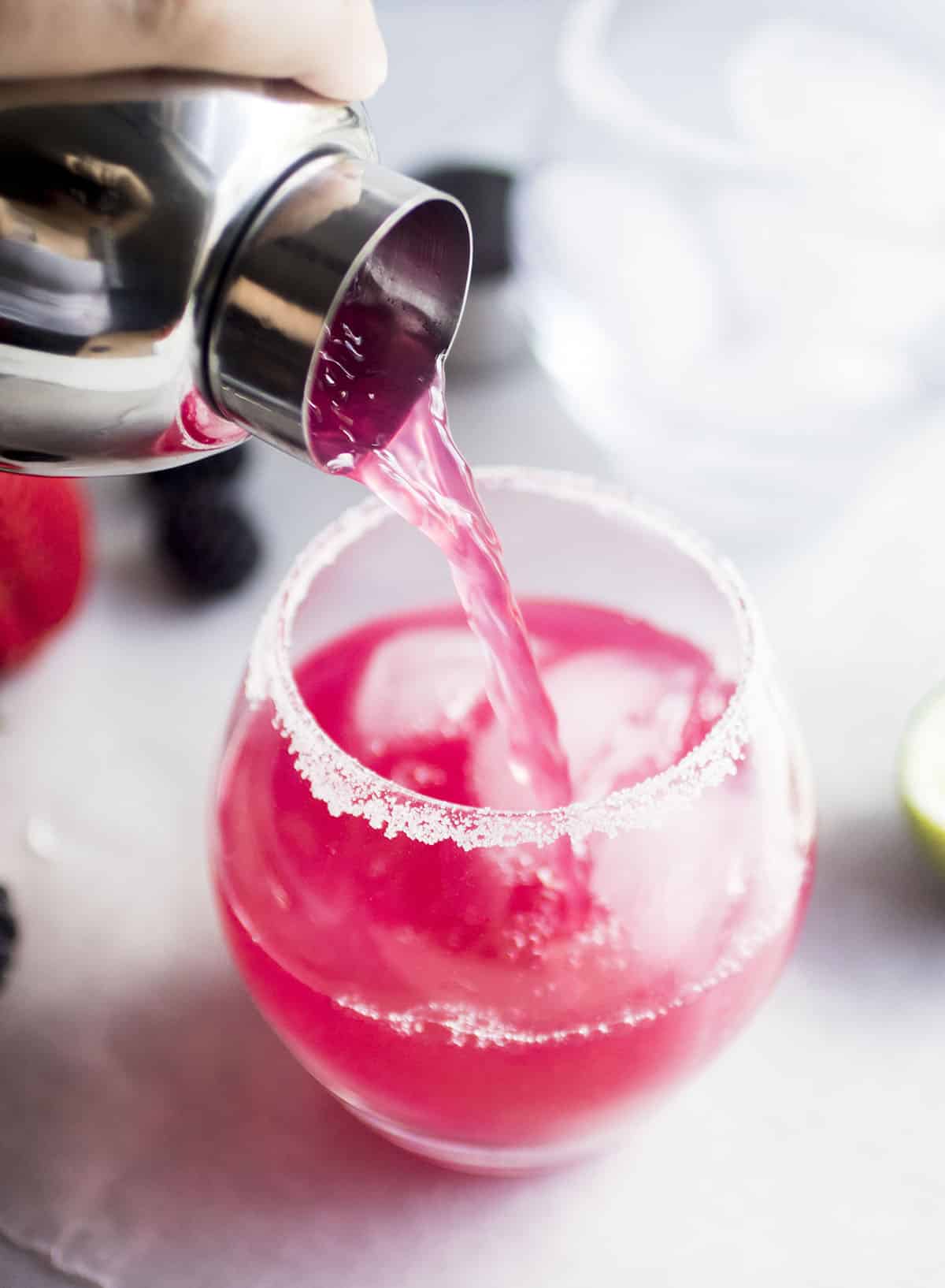 Pouring berry margaritas from a shaker into a prepared glass.