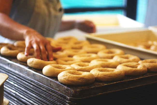 Hands placing formed bagels on a dark baking sheet.