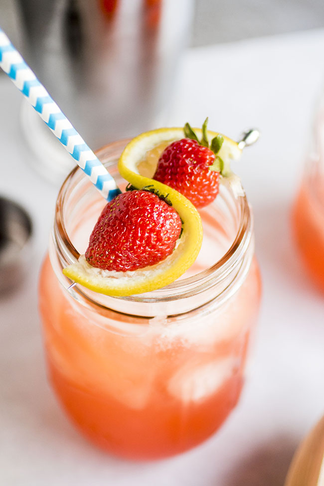 Cocktail in a mason jar with a blue striped straw, topped with whole strawberries and a slice of lemon.