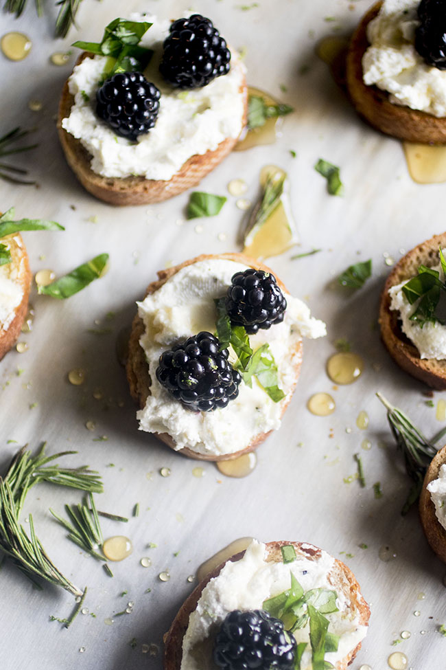 Crostini on a baking sheet lined with parchment paper, surrounded by fresh herbs and drops of honey.