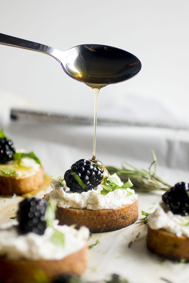 Spoon drizzling honey over crostini topped with goat cheese, blackberries, and basil.