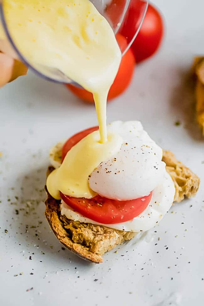 Clear measuring cup pouring hollandaise sauce over toast topped with tomato and a poached egg.