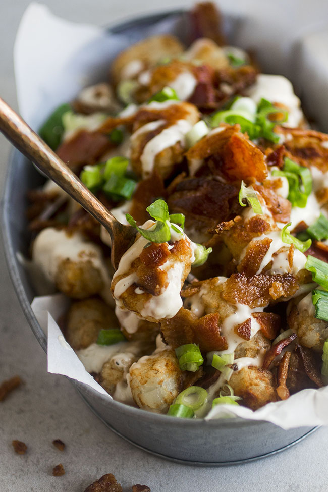 Bronze fork pulling a tater tot topped with bacon and green onions out of a metal serving dish.