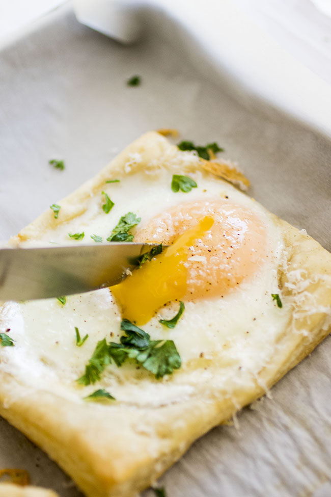 A knife cutting into the yolk of baked egg on a puff pastry square.