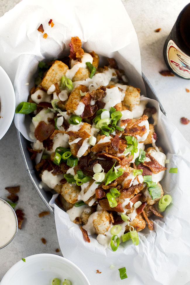 Tater tots topped with cheese sauce, bacon, and green onions on a white background.