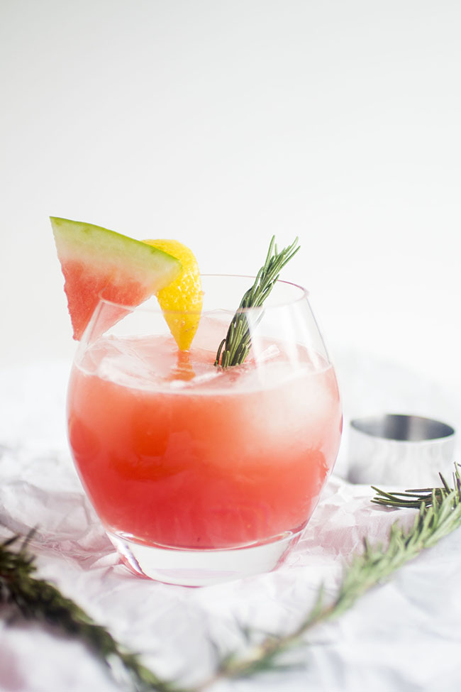 Watermelon cocktail garnished with a watermelon slice, a lemon wedge, and a sprig of fresh rosemary.