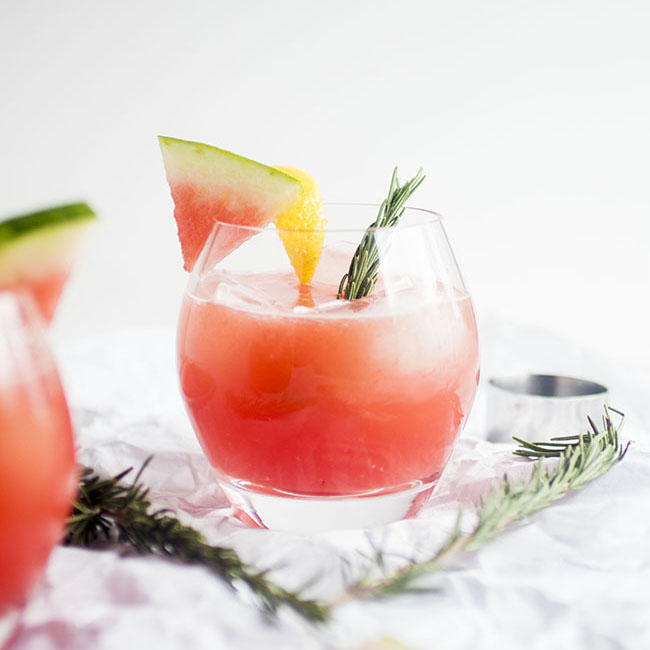 Pink cocktail garnished with a watermelon slice and a rosemary sprig.