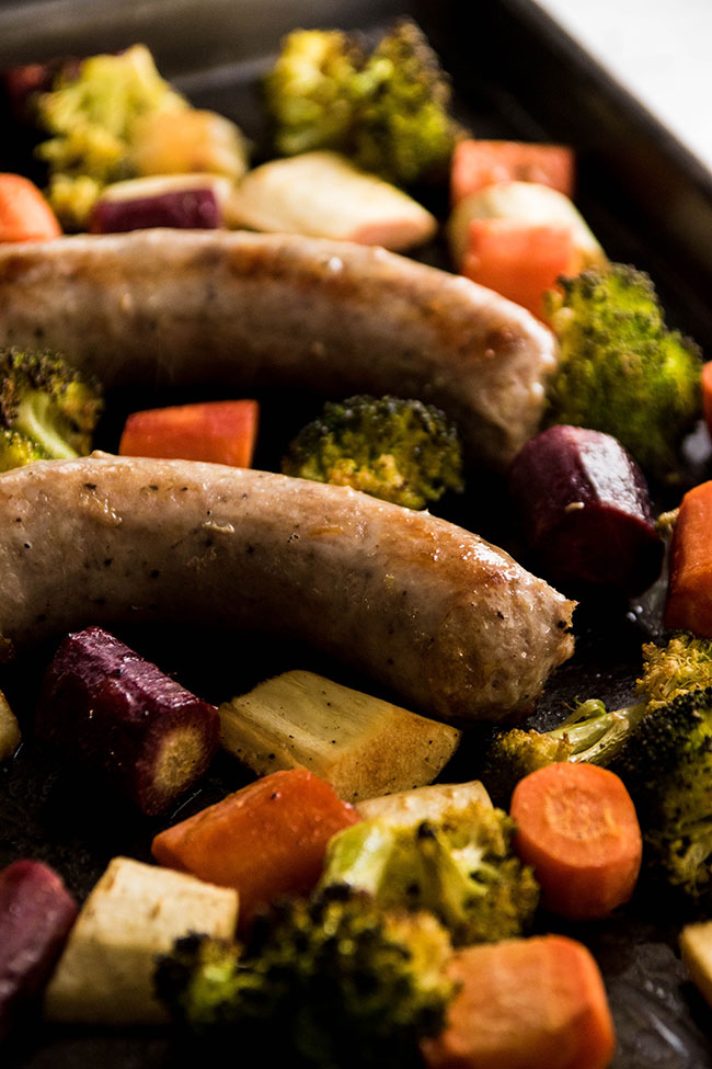 Close-up of cooked polish sausage next to chopped carrots, parsnips, and broccoli on a dark sheet pan.