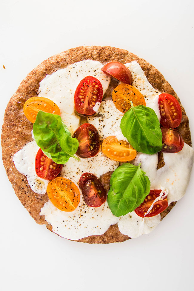 Wheat pita bread on a white background, topped with mozzarella cheese, cherry tomatoes, and fresh basil.
