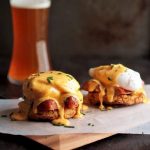 Two eggs benedict servings on a wooden cutting board in front of a glass of beer.