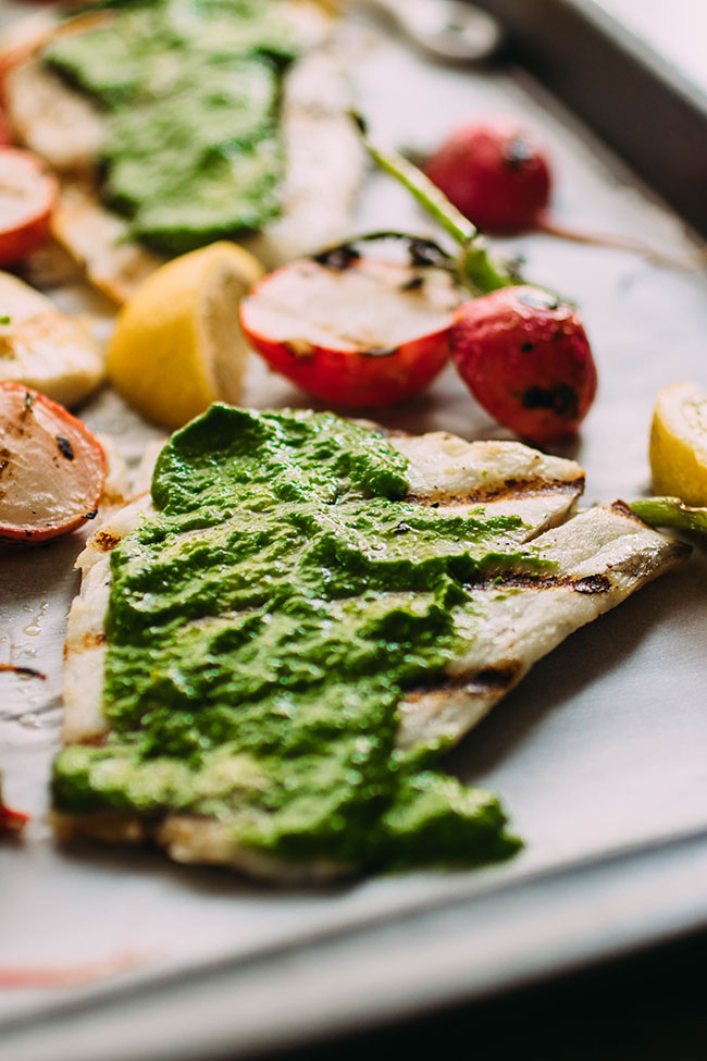 Barramundi fish fillet on a sheet pan, topped with bright green pesto with grilled radishes in the background.