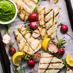 Grilled barramundi on a sheet pan with radishes, lemon wedges, and a bowl of pesto.