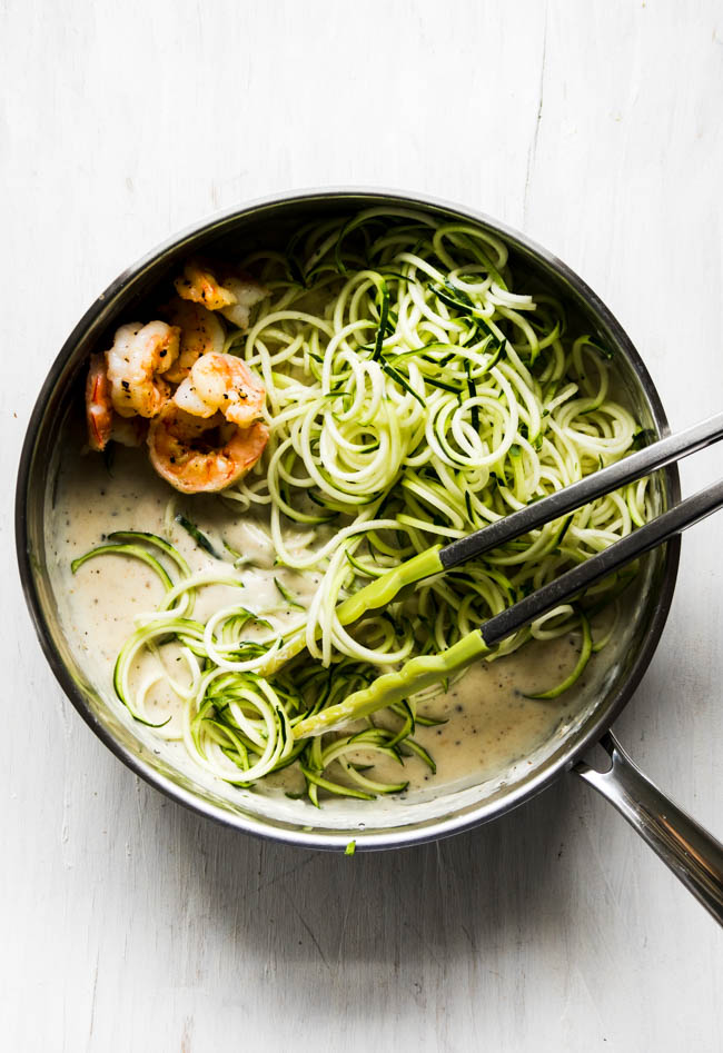 Tongs mixing zucchini noodles with alfredo sauce and shrimp in a large skillet.