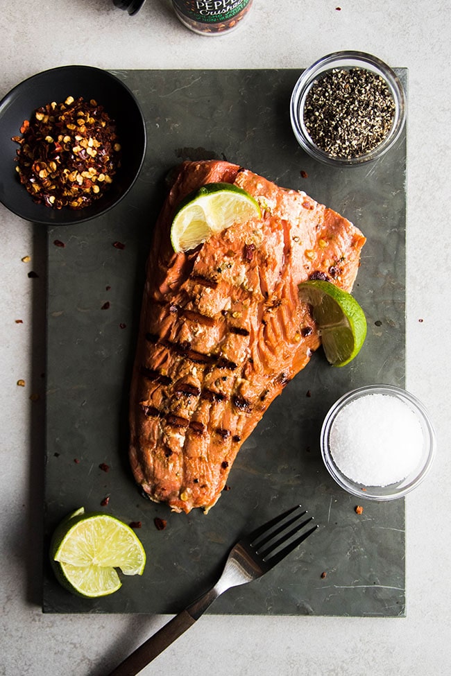 Grilled salmon fillet on a slate serving plate with small bowls of spices and several lime wedges.