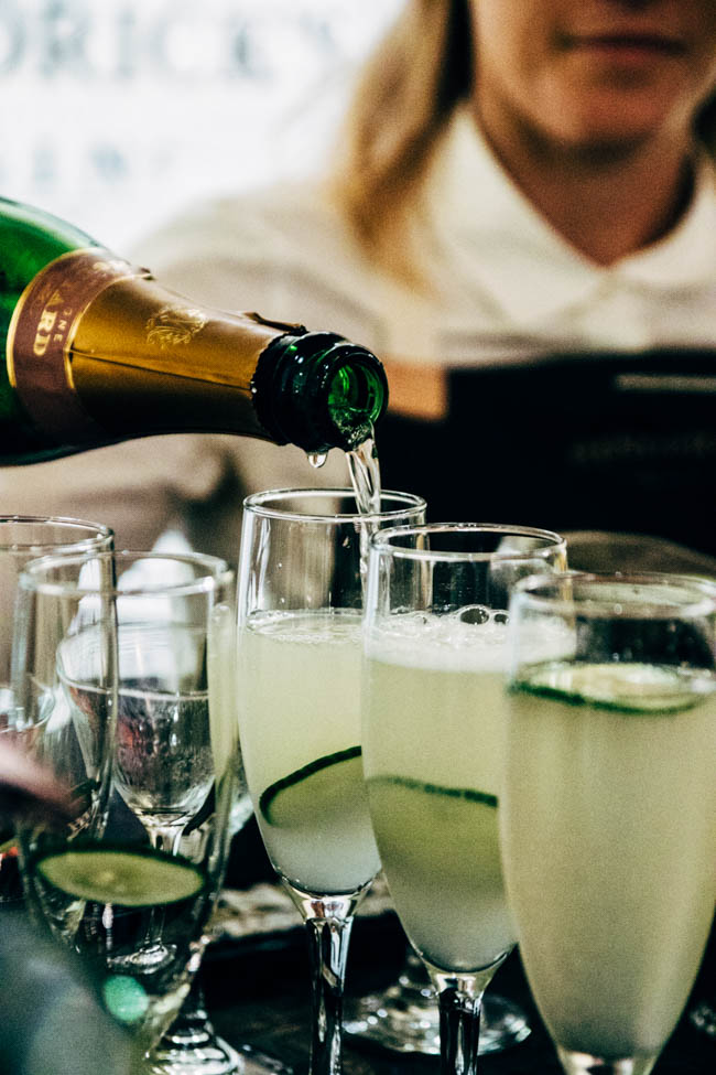 Prosecco being poured into champagne glasses with cucumber slices as a garnish.