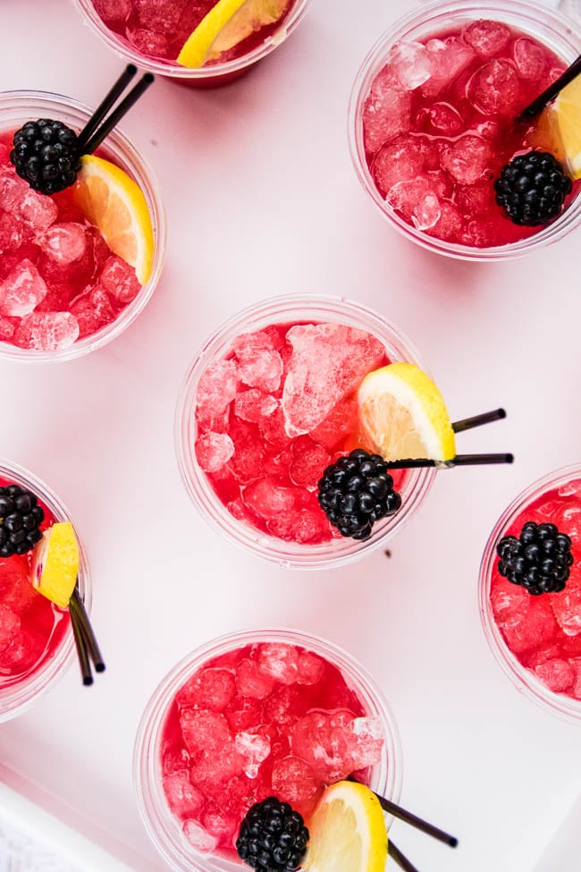 Aerial view of several plastic cups on a white table holding pink cocktails, garnished with fresh blackberries and lemon slices.