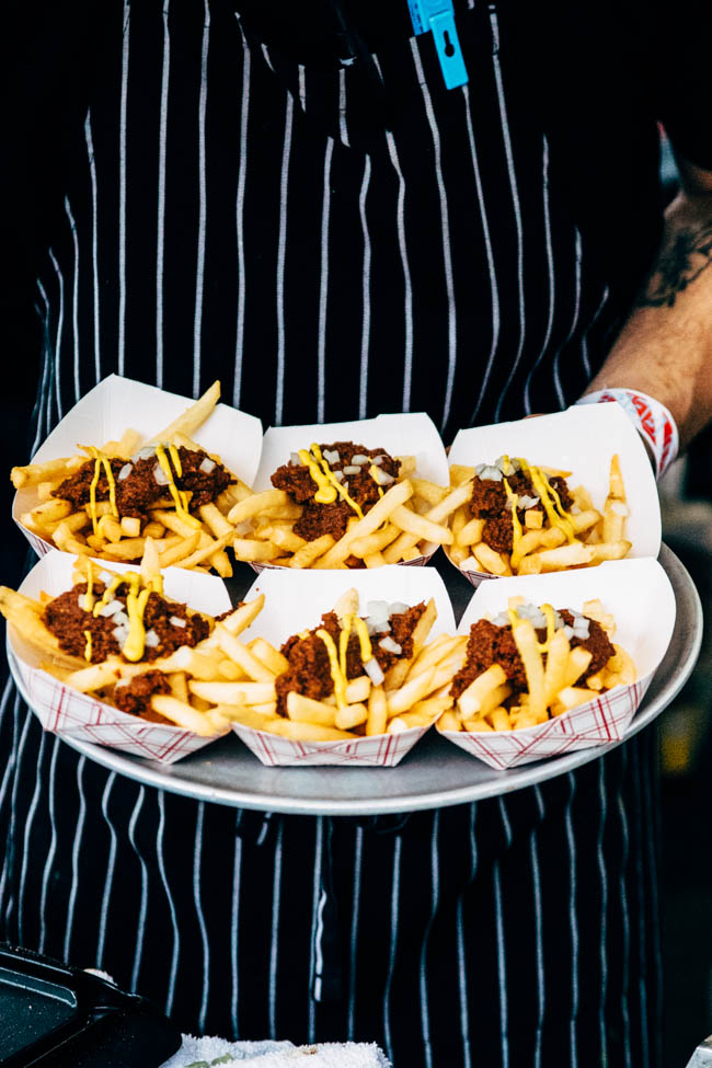 Hands holding a metal tray filled with individual portions of chili fries.