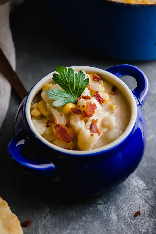 Chowder in a small blue bowl with handles, topped with chopped bacon and parsley on a dark background.