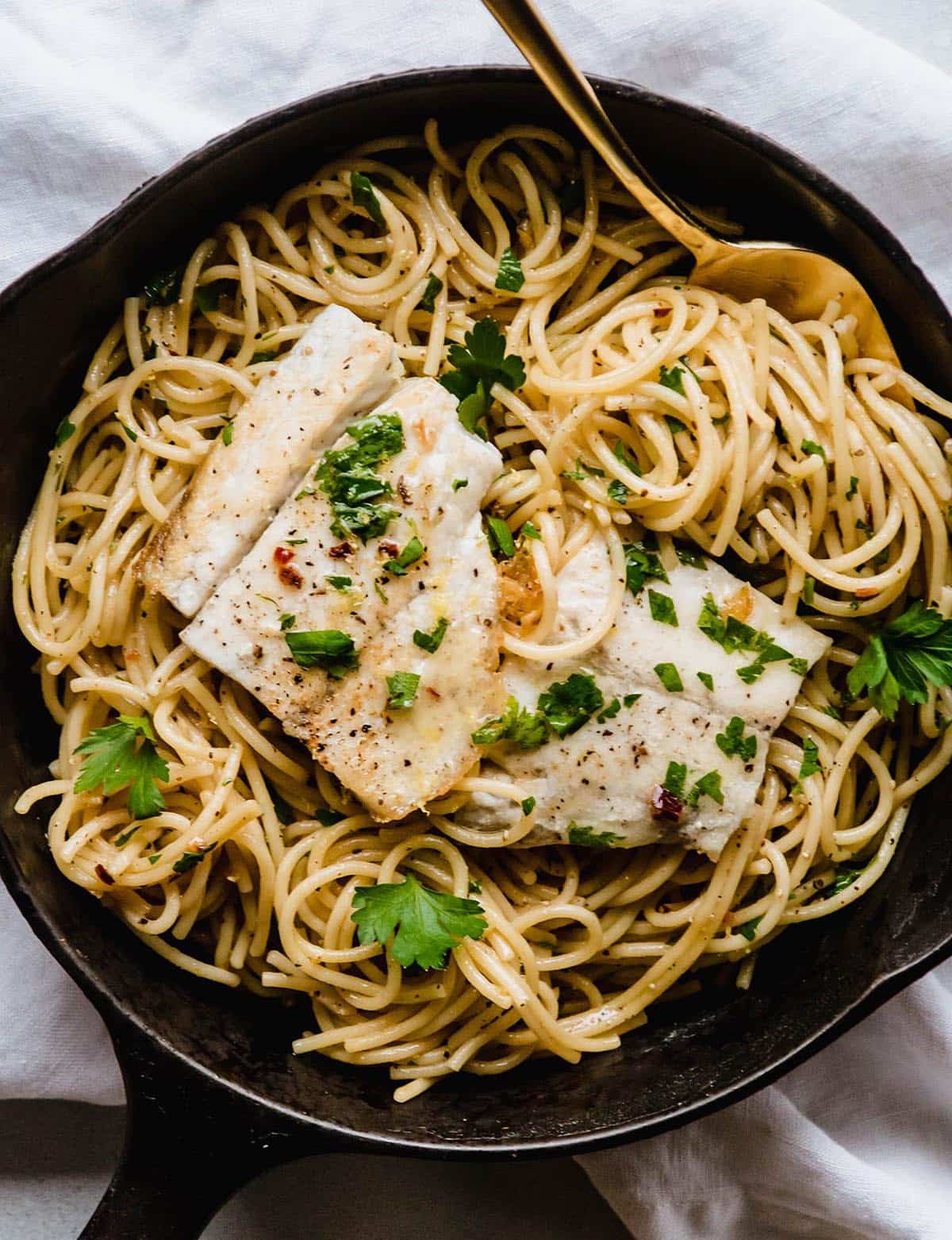 Large spoon twirling spaghetti in a cast iron skillet with cooked fish.