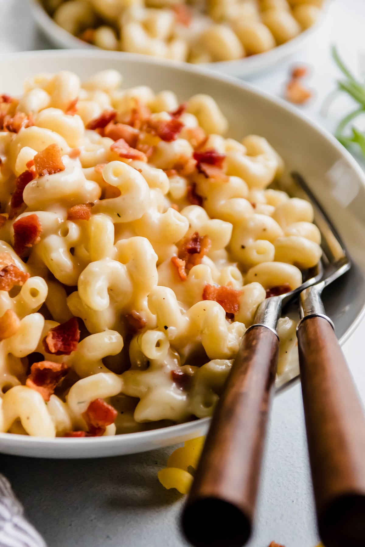 Two wooden forks in a large white bowl filled with macaroni and cheese and topped with crumbled bacon.