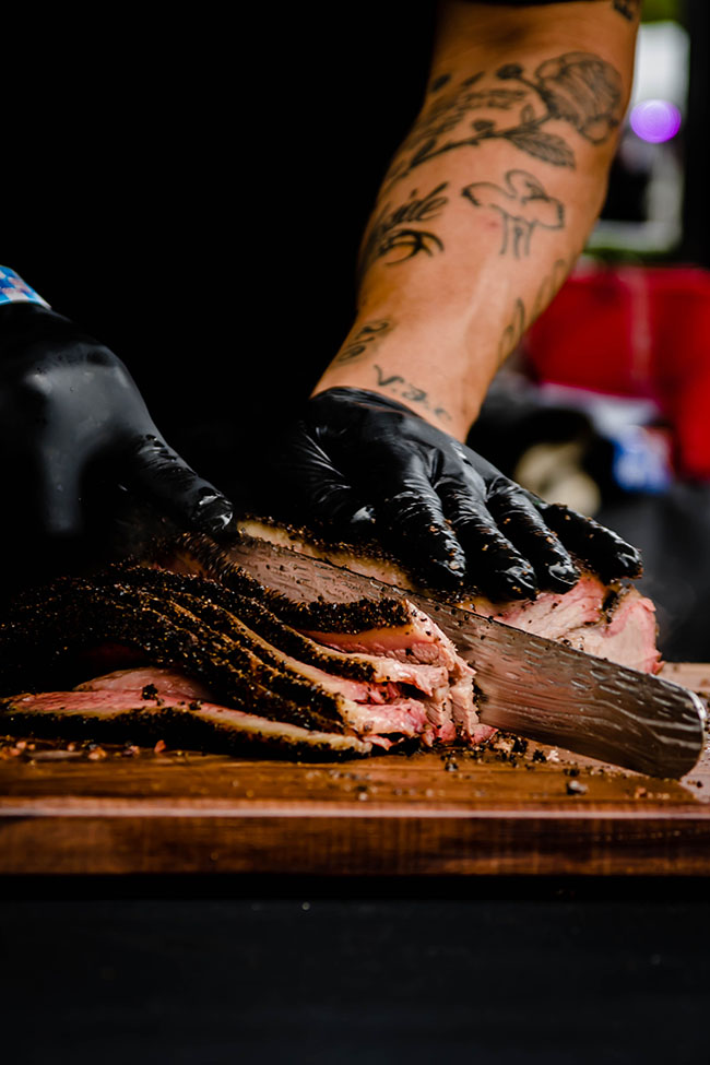 Hands in black gloves slicing a large piece of brisket on a wood cutting board.