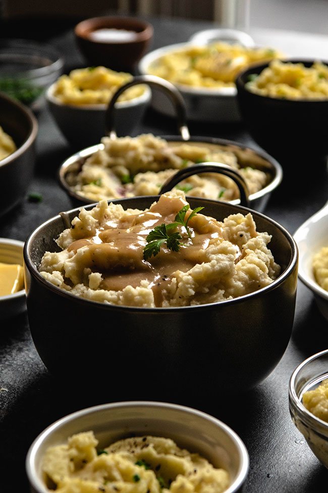 Mashed potatoes with gravy and parsley in a black bowl surrounded by multiple small bowls of potatoes and toppings.
