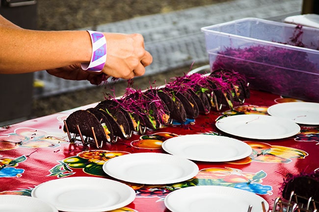 Hand putting a garnish on a row of tacos with purple shells.