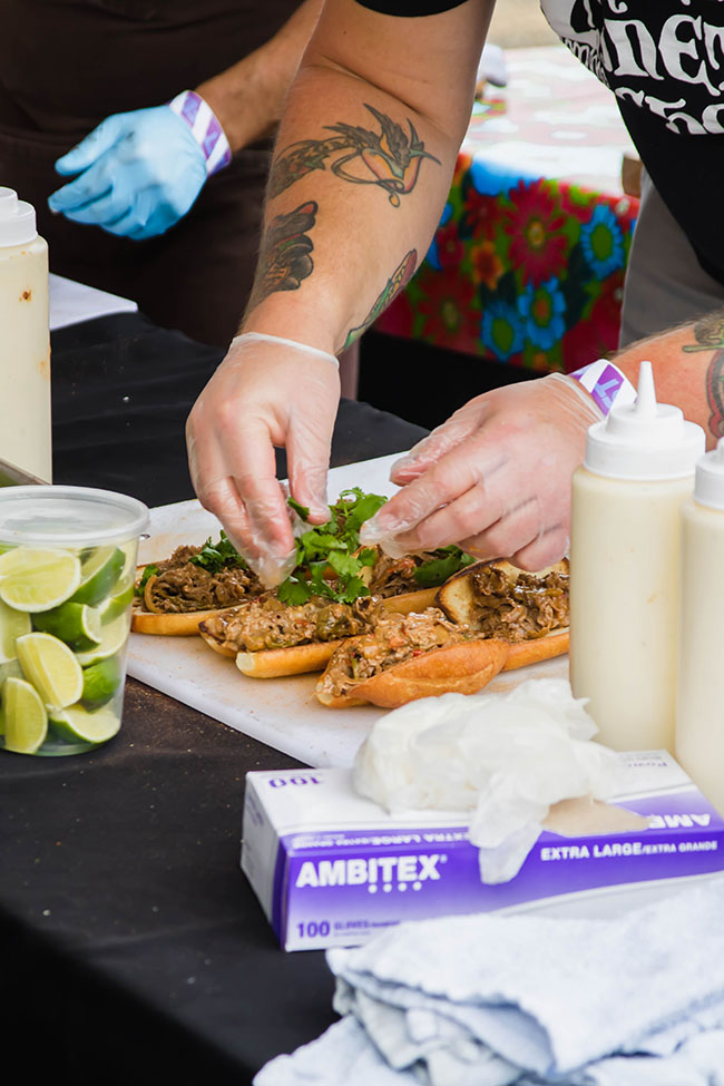 Man's hands assembling two banh mi sandwiches on a black prep table surrounded by ingredients.