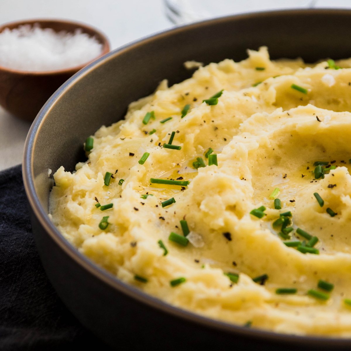 Mashed potatoes topped with fresh chives in a grey bowl on a white table.