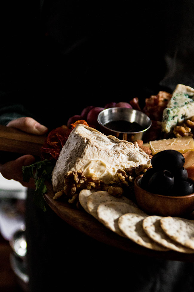 Woman in dark clothing holding a wooden cutting board filled with cheese and charcuterie.