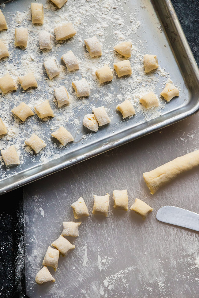 Sheet pan with fresh gnocchi next to a cutting board where gnocchi is being cut with a knife.