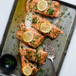 Baked steelhead fillet on a metal sheet pan, topped with fresh parsley and lemon slices and cut into portions.