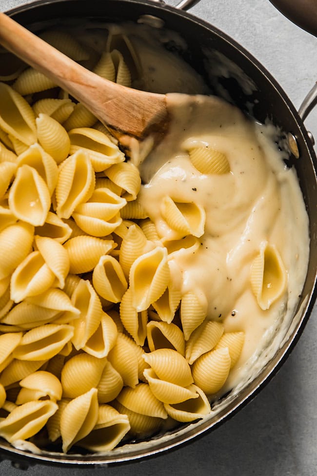 Wooden spoon stirring shell pasta into a pot of cheese sauce.