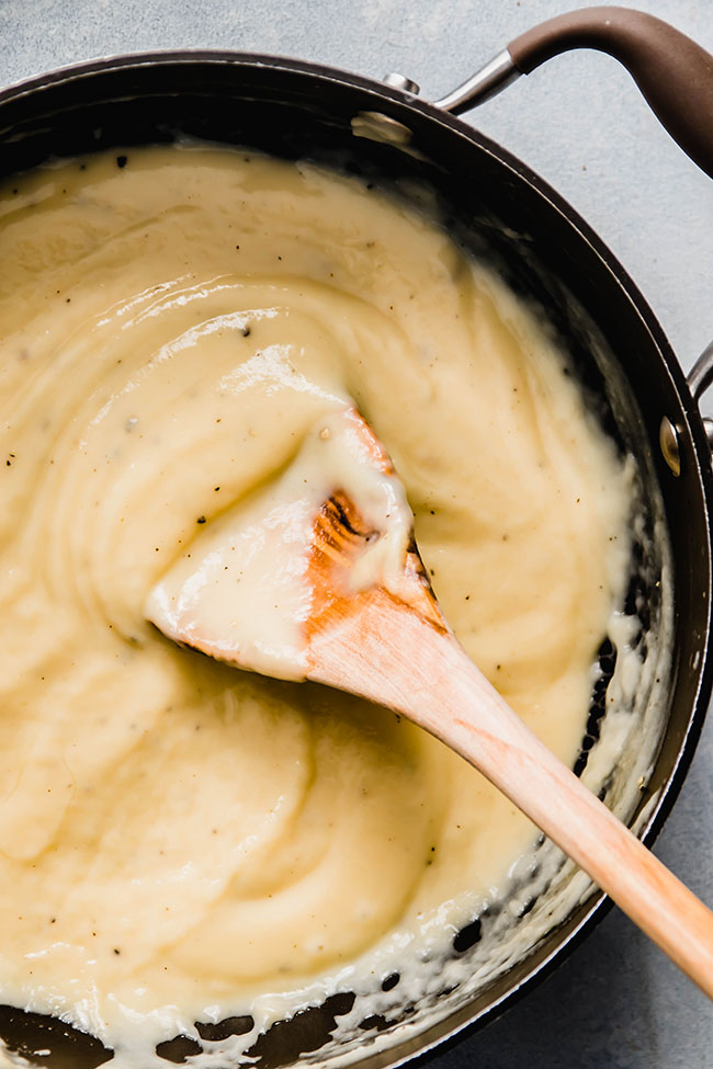 Wooden spoon stirring cheese sauce in a dark pot.