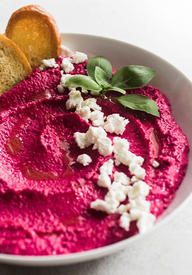 Pink beet dip in a white bowl, topped with crumbled goat cheese and fresh basil.