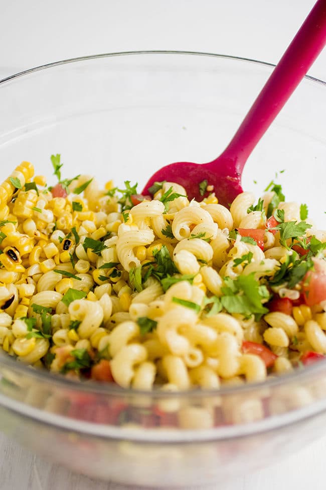 Red spatula stirring pasta salad together in a glass bowl.