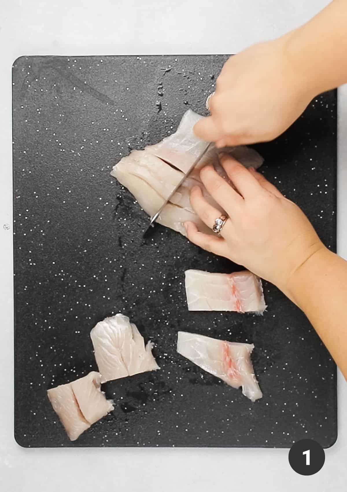 Hands cutting barramundi into small squares on a black cutting board.