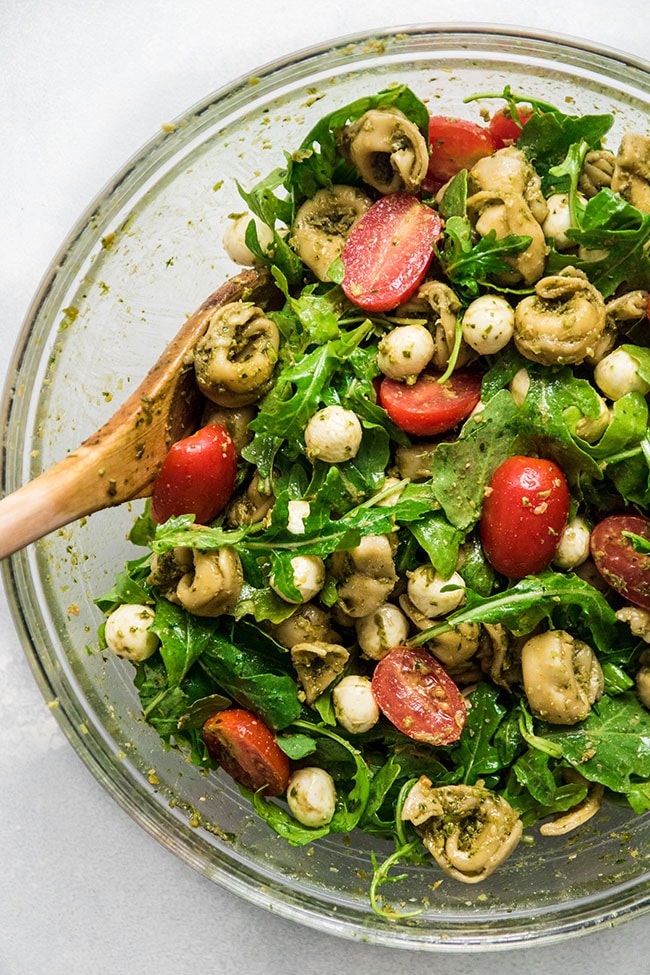 Tortellini salad with arugula and tomatoes in a glass bowl with a wooden spoon.