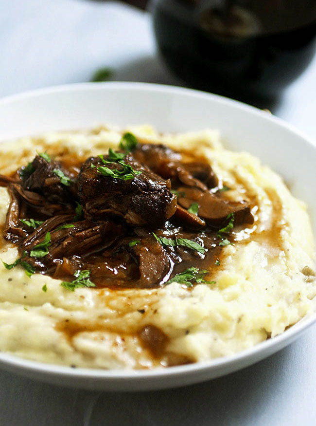 Braised beef short ribs on top of mashed potatoes in a shallow white bowl next to a glass of red wine.