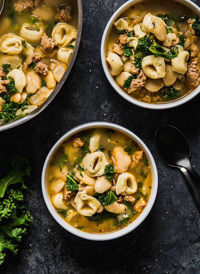 Two white bowls filled with tortellini soup on a black table.